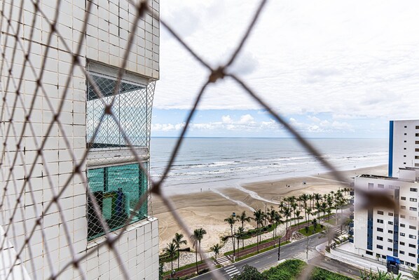 Une vue sur une plage, prise depuis un bâtiment dont la fenêtre est munie de barreaux, avec des palmiers et une route menant à la plage.