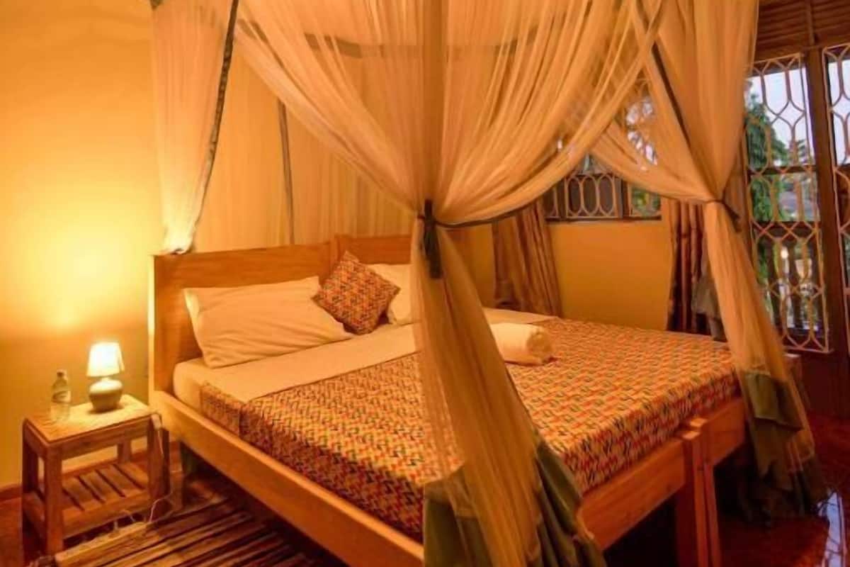 A room with a bed under a canopy, a nightstand with a lamp, and a window with a view.