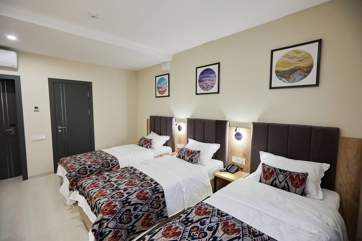 Habitación de hotel con tres camas, cada una con ropa de cama estampada y cabecero. Se observan obras de arte colgadas en la pared y una mesita de noche con lámpara.