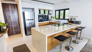 A modern kitchen with a large island, bar stools, a refrigerator, oven, and a window with a view of greenery.