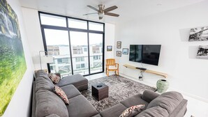 A modern living room with a large gray sectional sofa, a flat-screen TV mounted on the wall, and a large window offering a city view.