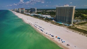 An aerial view of a beach with a row of hotels along the shore, featuring a large building with a flat roof and a smaller building with a curved roof.