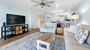 A spacious living room with a television, a ceiling fan, and a kitchen area in the background. - Ramsgate 31 - Gulf-Front Escape + Beach Service (Panama City Beach)
