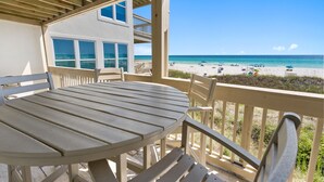 A covered outdoor area with a table and chairs, overlooking a beach with umbrellas and people. - Ramsgate 31 - Gulf-Front Escape + Beach Service (Panama City Beach)