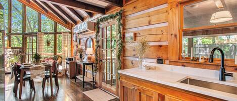 A wooden cabin interior with a dining area, kitchen, and large windows.