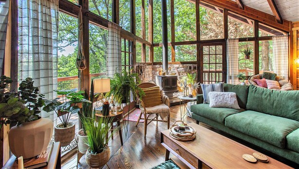 A spacious room with large windows, a green sofa, a wooden table, and various potted plants. - Chic Boho Cabin in the Trees! (Ellijay)