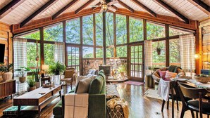 A spacious living room with a high ceiling, large windows, wooden beams, a fireplace, and a television. - Chic Boho Cabin in the Trees! (Ellijay)