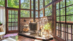 A cozy cabin interior with a stone fireplace, wooden beams, and large windows offering a view of the forest. - Chic Boho Cabin in the Trees! (Ellijay)