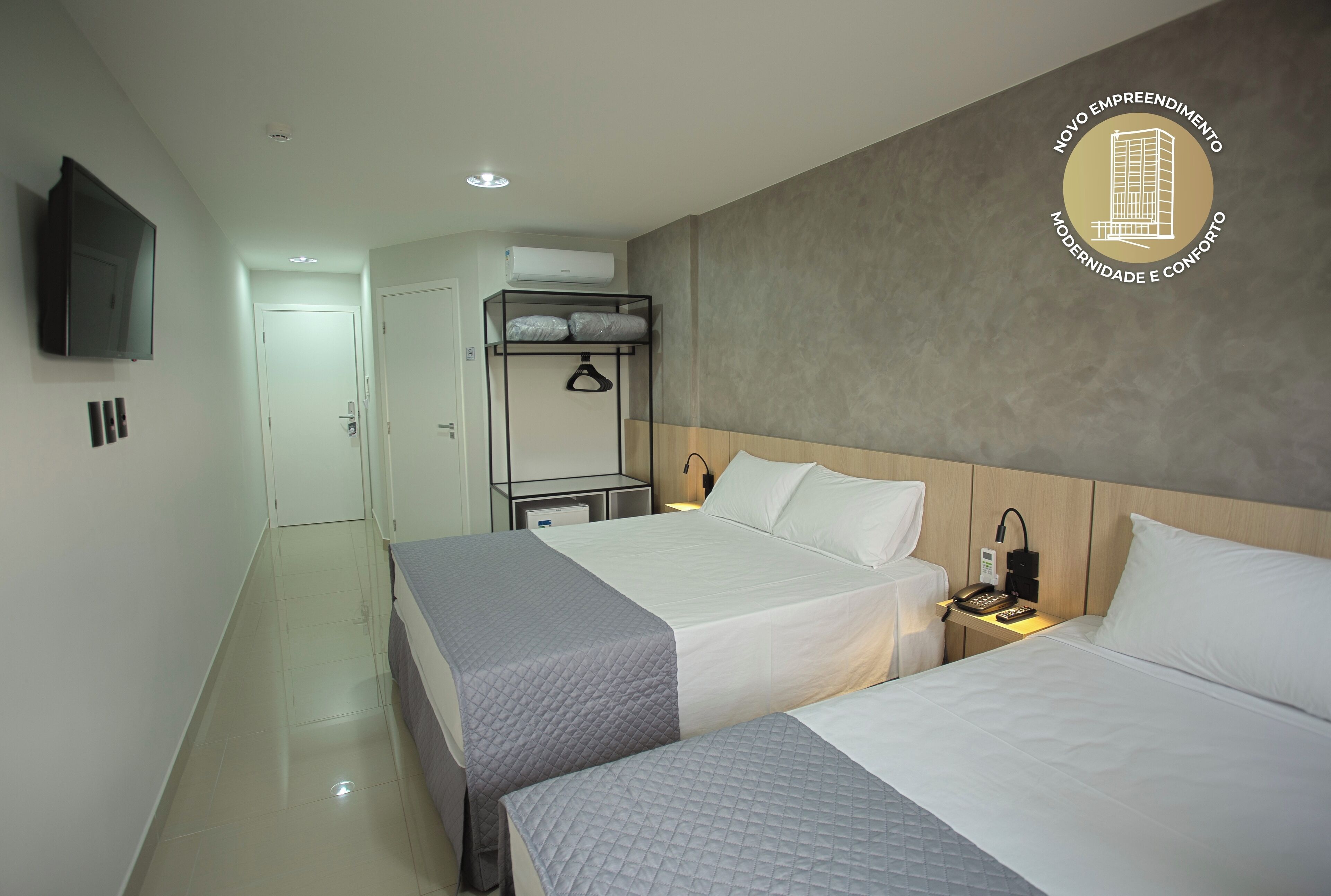 Quarto de hotel com duas camas, televisão, ar condicionado, mesa de cabeceira com luminária e guarda-roupa com cabides.