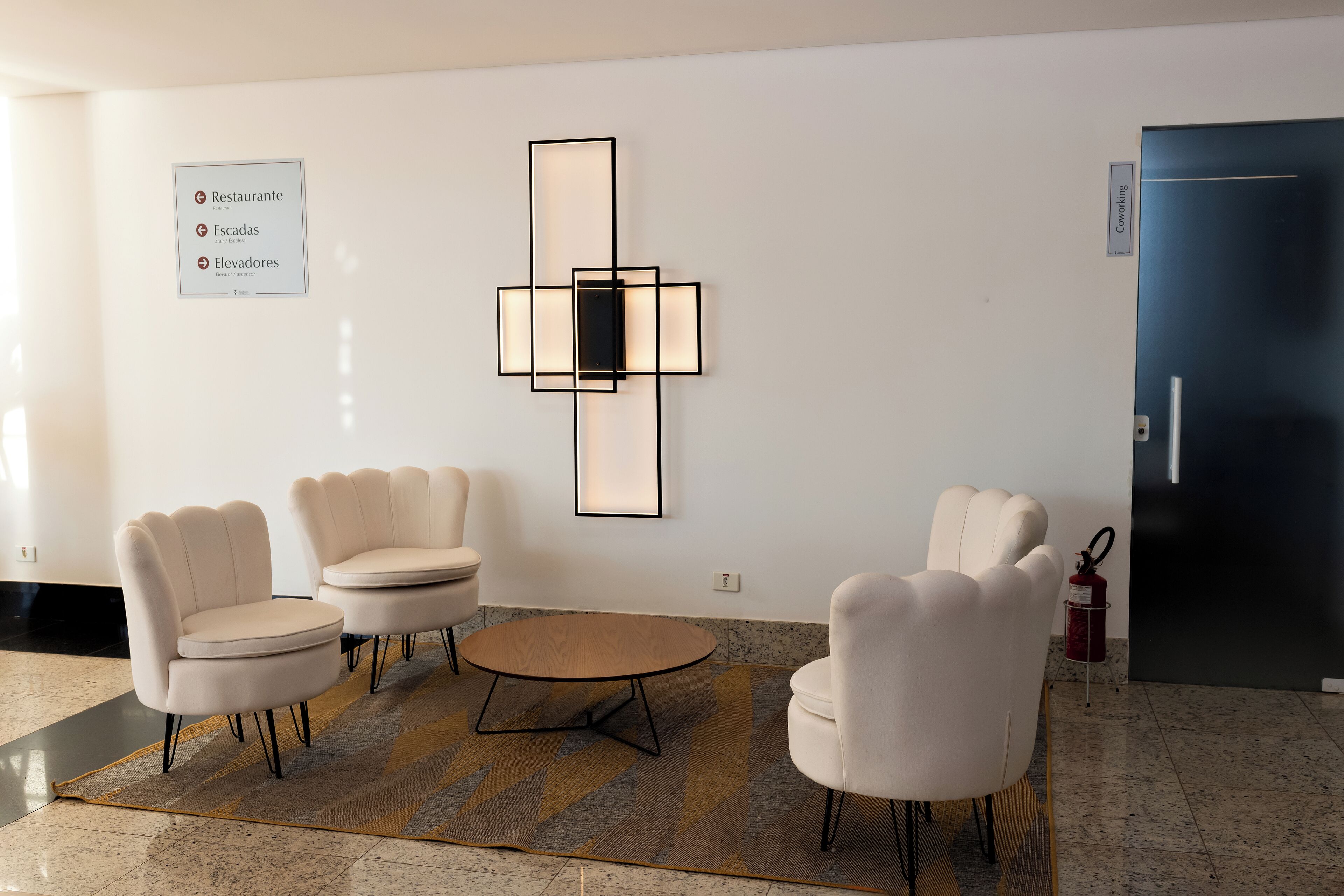 A modern lobby with a minimalist design, featuring a seating area with four chairs and a round table, a wall-mounted light fixture, and a fire extinguisher.