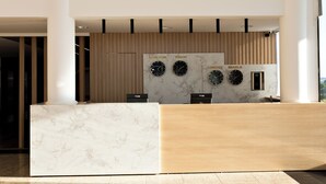 A reception desk with a marble top and a wooden panel wall behind it. There are clocks displaying different time zones and the words "Nova York," "Toquio," "Londres," and "Brasilia" on the wall. - GoldMen Express Cianorte (Cianorte)