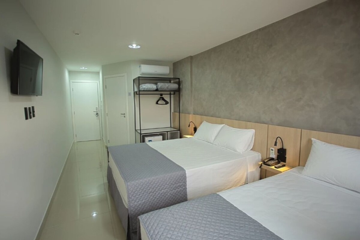 Quarto de hotel com duas camas, um guarda-roupa, uma televisão e uma mesa de cabeceira com abajur.