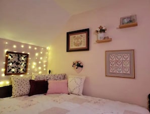 A bedroom with a bed, decorative wall lights, a framed picture, and a small shelf with flowers.