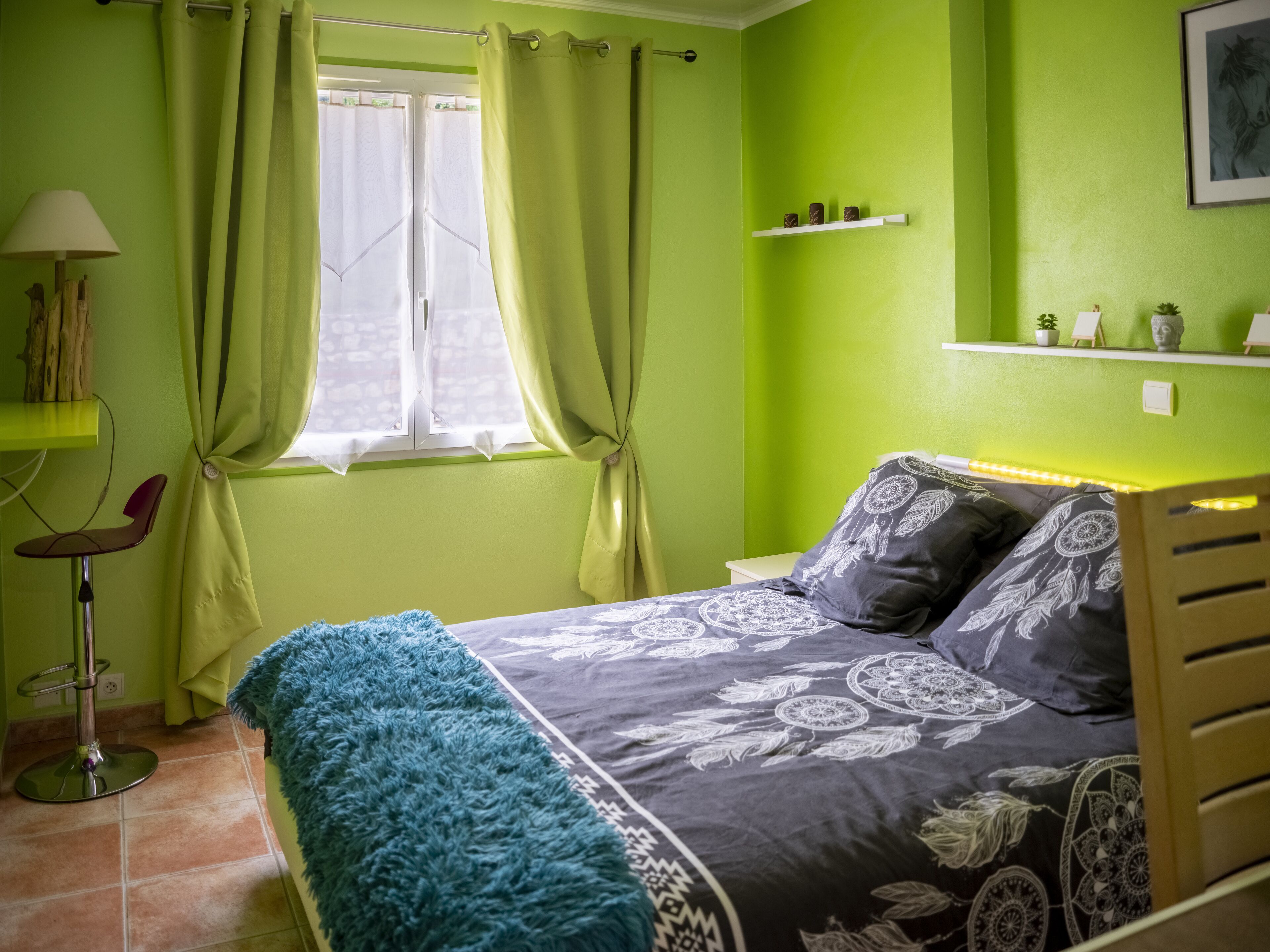 A bedroom with a bed, a bedside table, a lamp, a window with curtains, a shelf with decorative items, and a wall with a framed picture.