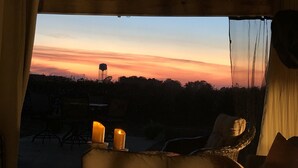 A room with a view of a sunset over a water tower, a wicker chair, and two lit candles on a table. - Sunset Log Cabin (Clarksdale)