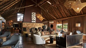 A spacious lounge area with a high ceiling, rustic wooden beams, and a stone fireplace. There are multiple seating options including sofas and armchairs. - Letamo at Qwabi Private Game Reserve by Newmark (Thabazimbi)