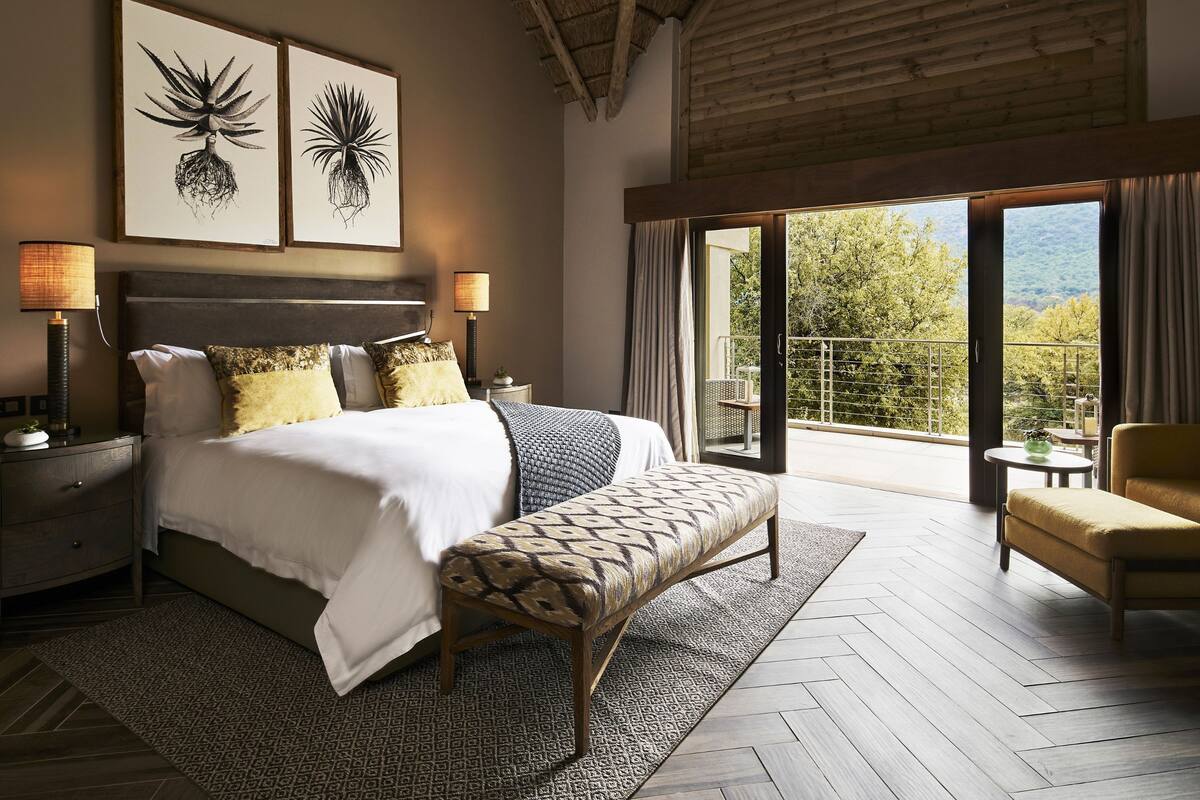 Une chambre avec un grand lit, deux tables de chevet avec des lampes, un banc et un balcon donnant sur la forêt.