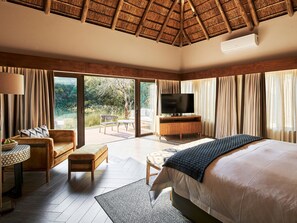 A bedroom with a large bed, a TV, a wooden cabinet, and a seating area with a table and chairs. - Letamo at Qwabi Private Game Reserve by Newmark (Thabazimbi)