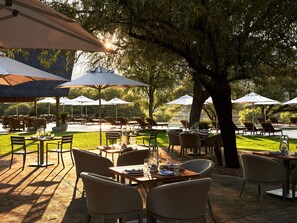 An outdoor dining area with tables and chairs, umbrellas, and a view of a body of water. - Letamo at Qwabi Private Game Reserve by Newmark (Thabazimbi)