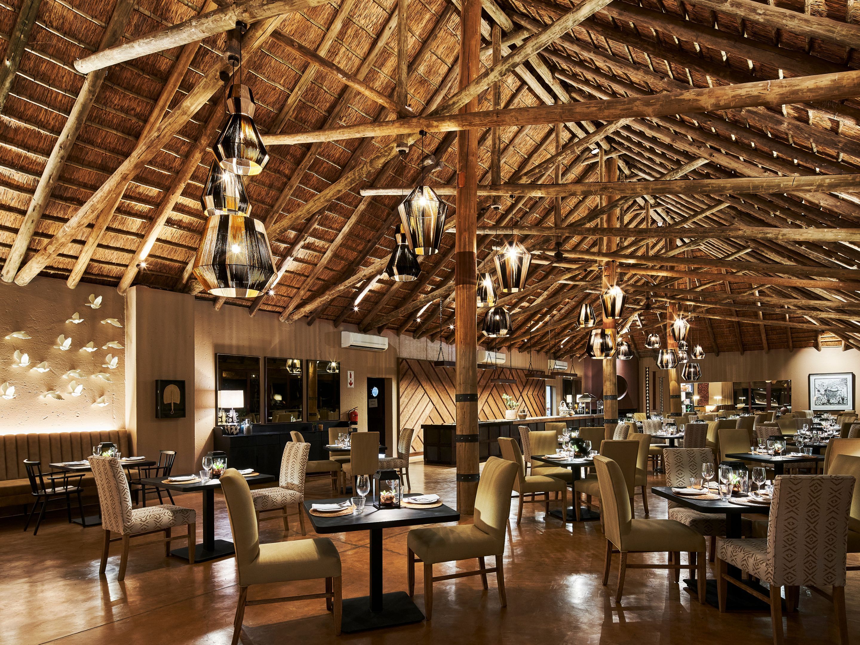 Un restaurante espacioso con techos altos de madera y lámparas colgantes. Las mesas están dispuestas con sillas, y el piso es de madera.