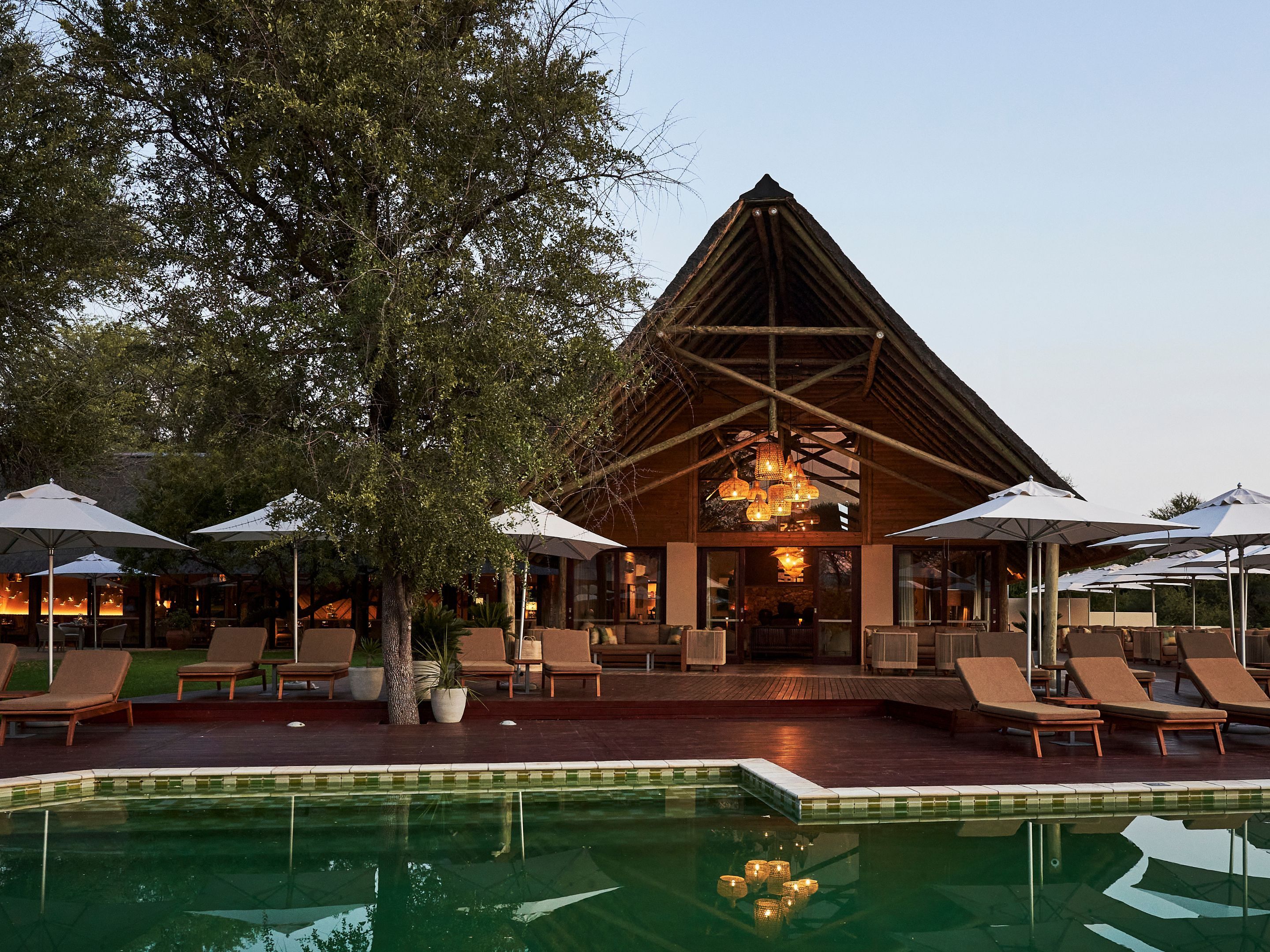 Un espace aménagé au bord de la piscine avec des chaises longues et des parasols, un bâtiment en bois doté d’un toit triangulaire et un luminaire suspendu.