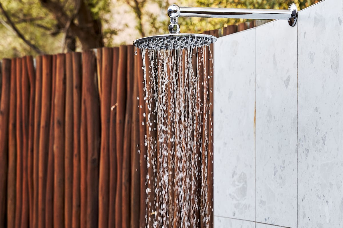 Une douche avec de l’eau qui coule le long d’une barre chromée, une clôture en bois en arrière-plan et un mur recouvert de carreaux blancs.