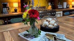 A wooden kitchen table with a floral centerpiece, a glass dome with cookies, and a blue polka-dotted dish.