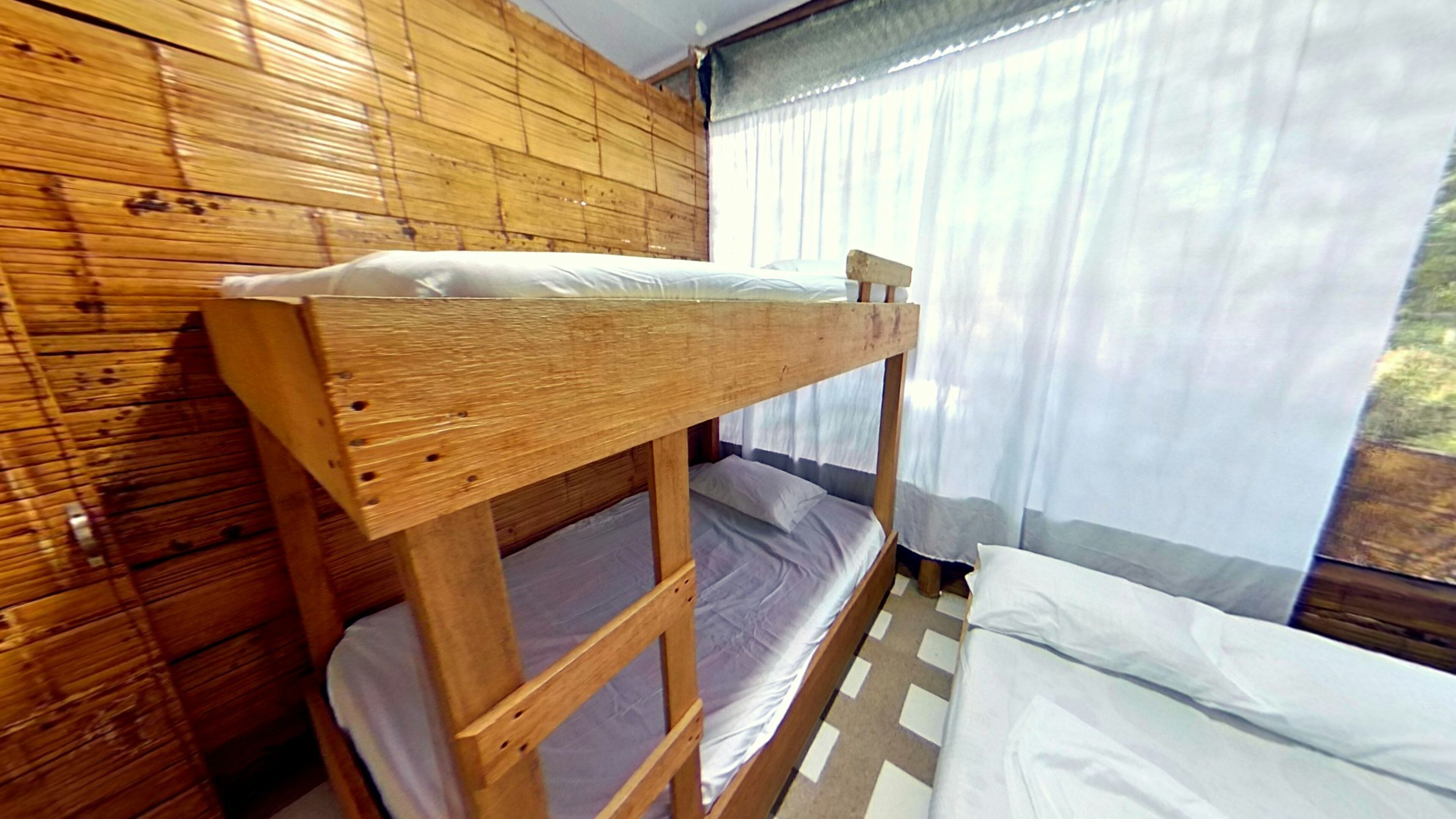 A bunk bed in a room with wooden walls and a window with sheer curtains.