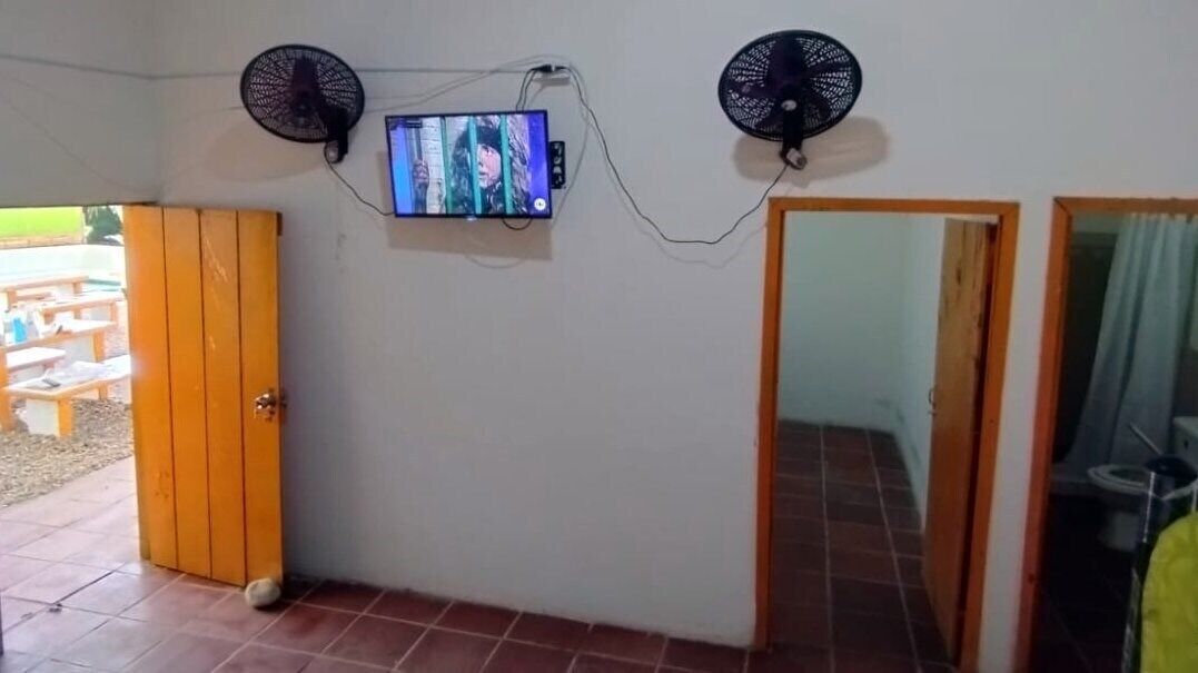 A room with a television mounted on the wall, two wall fans, a wooden door, and a tiled floor.
