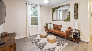 A living room with a brown leather sofa, two round ottomans, a ceiling fan, a large mirror with a green frame, and a small side table with a lamp. - Quiet get away in a vibrant city. (St. Augustine)