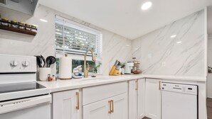 A modern kitchen with white cabinetry, a built-in oven, a dishwasher, and a window with blinds. - Quiet get away in a vibrant city. (St. Augustine)