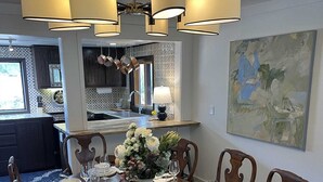 Zona pranzo con un tavolo rotondo apparecchiato per sei persone, circondato da sedie in legno. Sopra, un lampadario e sul tavolo un vaso con fiori.