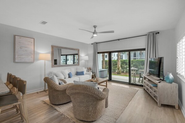 A modern living room with a white sofa, two woven chairs, a coffee table, and a TV on a wooden stand. - Beachside 30A Condo with Private Beach (Santa Rosa Beach)