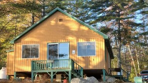 A wooden cabin with a green porch and two windows, surrounded by trees. - Beautiful lakefront cottage, Ambajejus Lake (Northern Piscatiquis Cnt)