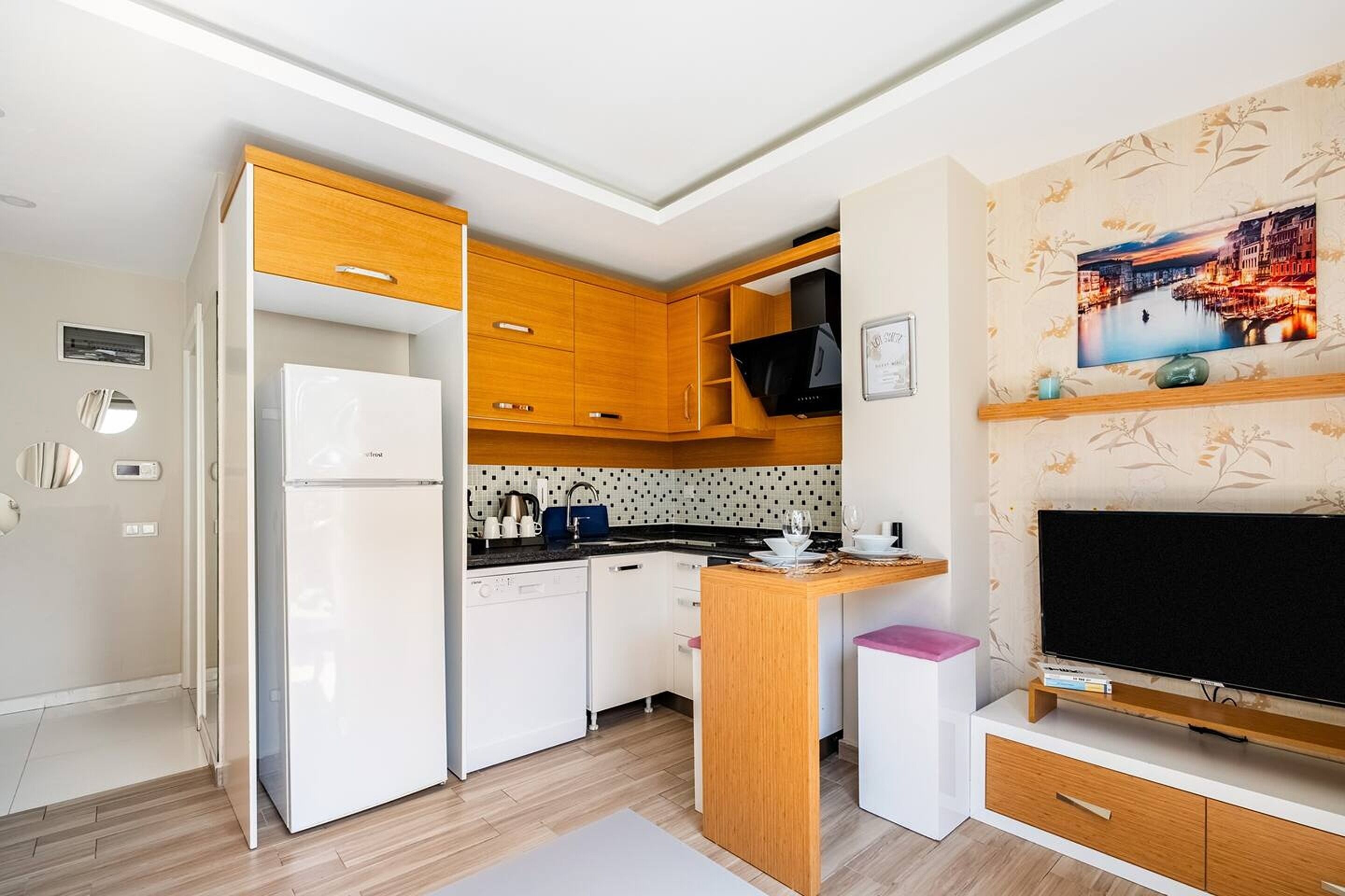 Een kleine keuken met een witte koelkast, houten keukenkastjes, een magnetron en een zwart fornuis. Er is een zithoekje met een tafel en stoelen, en een flatscreen televisie op een houten standaard.