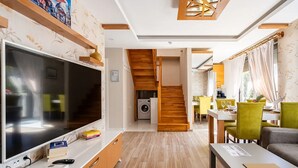 A modern living room with a large flat-screen TV, wooden flooring, a staircase, and a dining area with yellow chairs. - Loi Suite (Antalya)