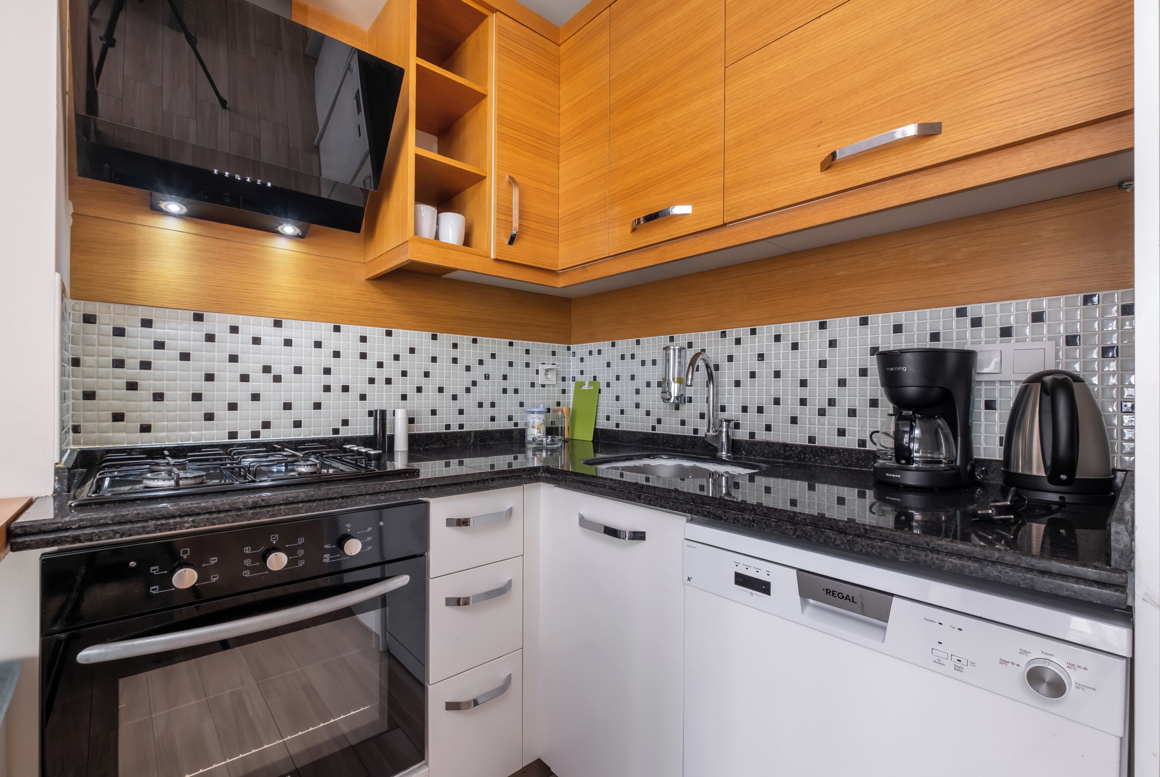 A modern kitchen with wooden cabinets, a black countertop, a gas stove, a dishwasher, and a tiled backsplash.