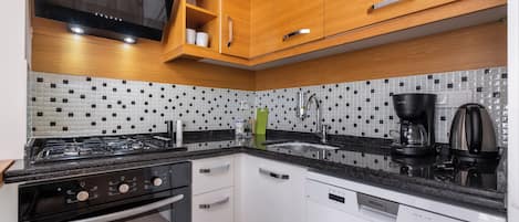 A modern kitchen with wooden cabinets, a black countertop, a gas stove, a dishwasher, and a tiled backsplash.
