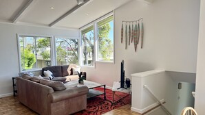 Amplia sala de estar con un sofá grande, una mesa de centro y un televisor. Amplias ventanas ofrecen luz natural y vistas a la vegetación exterior.