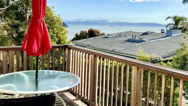 Una terraza de madera con una mesa de vidrio y un paraguas rojo, con vista a un puerto deportivo con barcos y una vista distante del horizonte de la ciudad.
