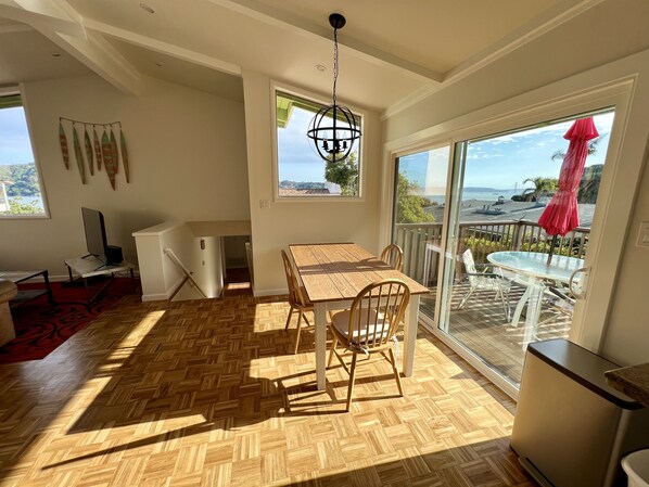 A spacious room with a wooden herringbone floor, a dining table with chairs, a large window with a view of the sea, and a balcony with outdoor furniture. - Stay near San Francisco Bay, enjoy our 2+bedroom Tiburon retreat (Tiburon)