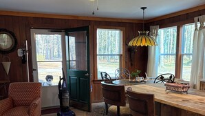 Interior acollidor d'una cabana amb una zona de menjador de fusta, un ventilador de sostre i grans finestres que donen vista al bosc.