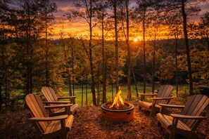 A campfire with four Adirondack chairs around it, set in a forested area with a sunset view. - Golf View located at Innsbruck Resort! Minutes to Oktoberfest! (Helen)