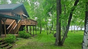 A wooden cabin with a porch and a deck, surrounded by lush greenery and trees.