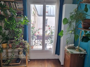 A room with a bookshelf, a potted plant, a window with a view of a balcony, and a desk with a lamp.