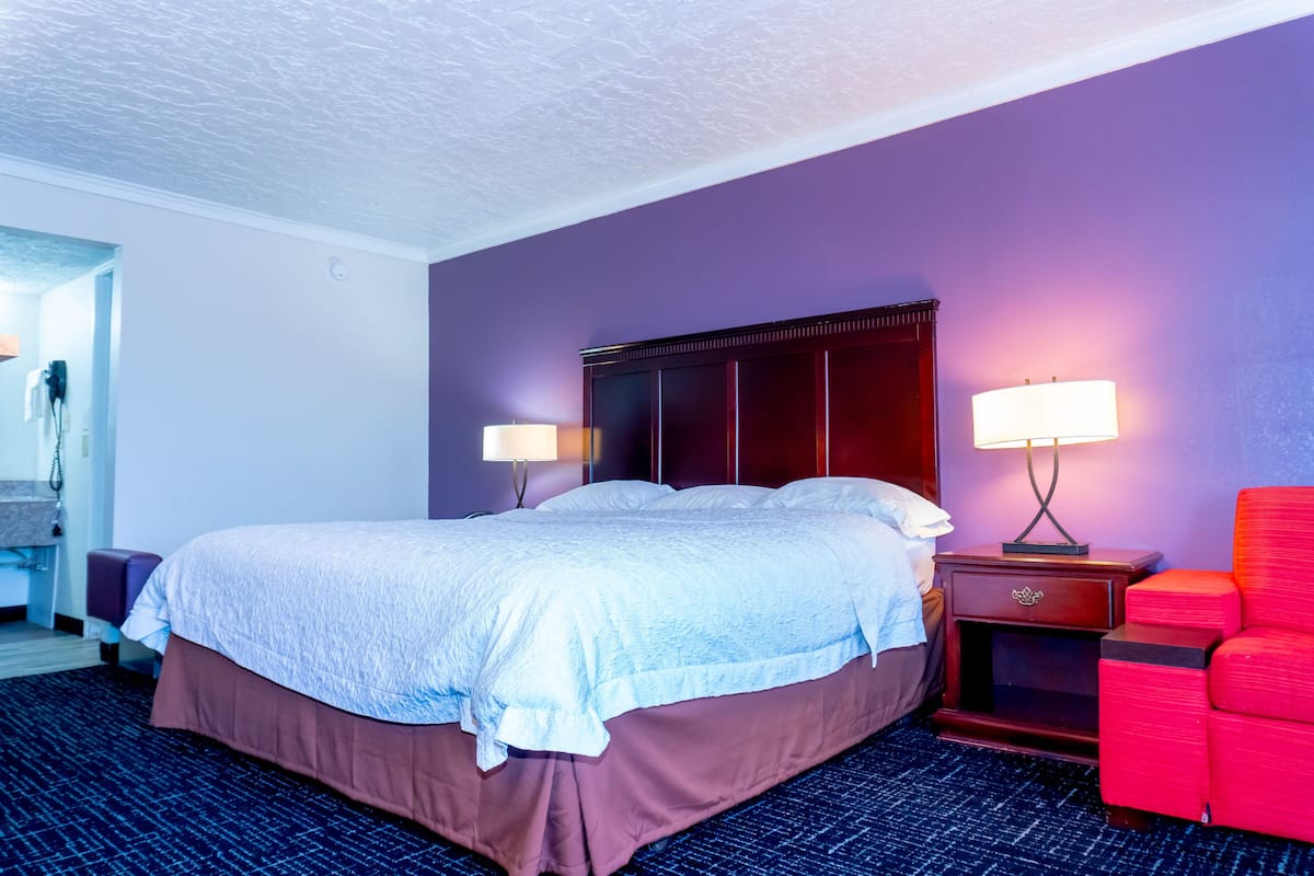 A hotel room with a large bed, two bedside lamps, a red chair, and a wooden nightstand.