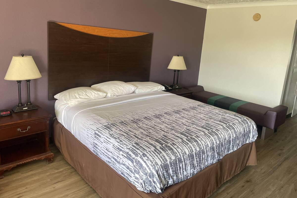 A hotel room with a large bed, two bedside tables with lamps, a wooden headboard, and a bench by the door.