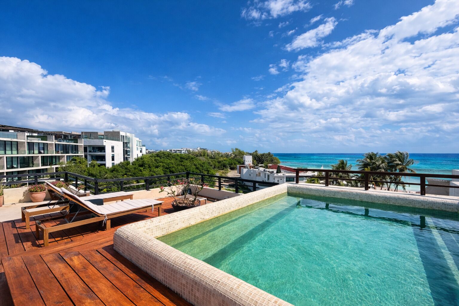 Une piscine sur le toit, avec une eau d’un bleu limpide, une terrasse en bois, des chaises longues et une vue sur l’océan et les bâtiments.
