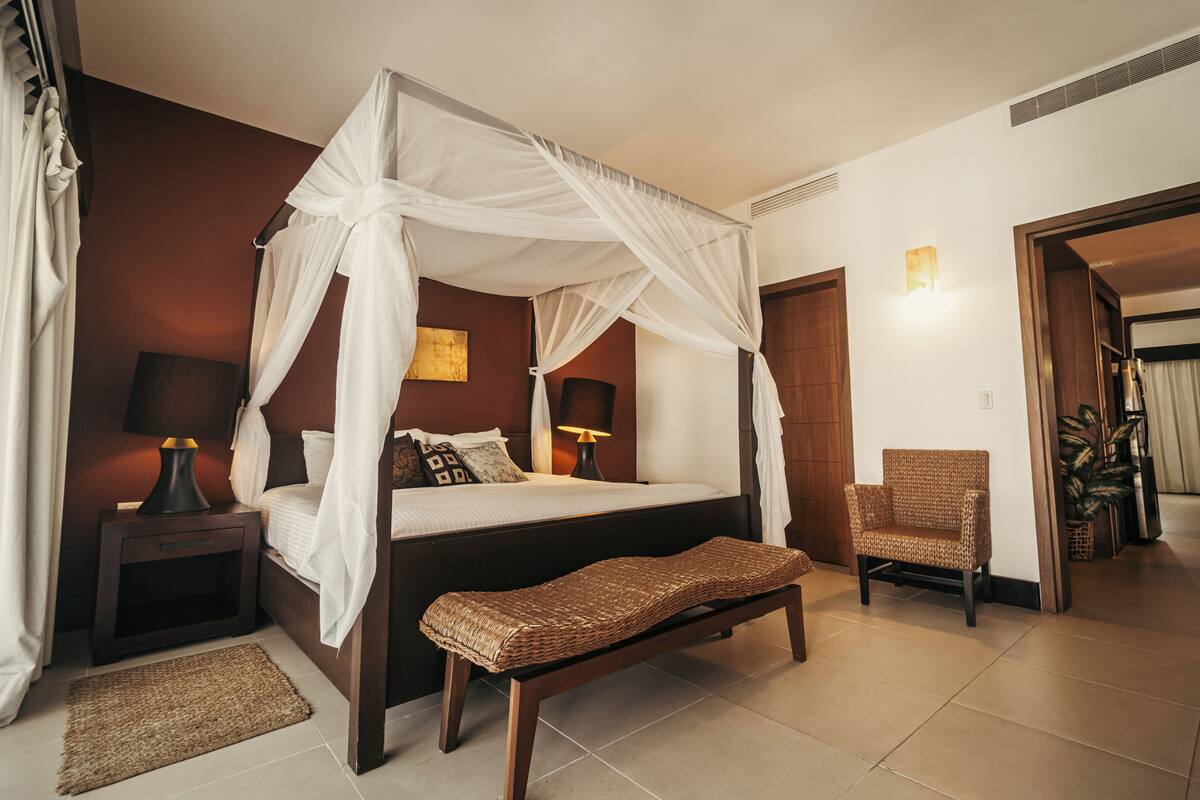 A bedroom with a four-poster bed, bedside tables, a bench, a chair, and a lamp.