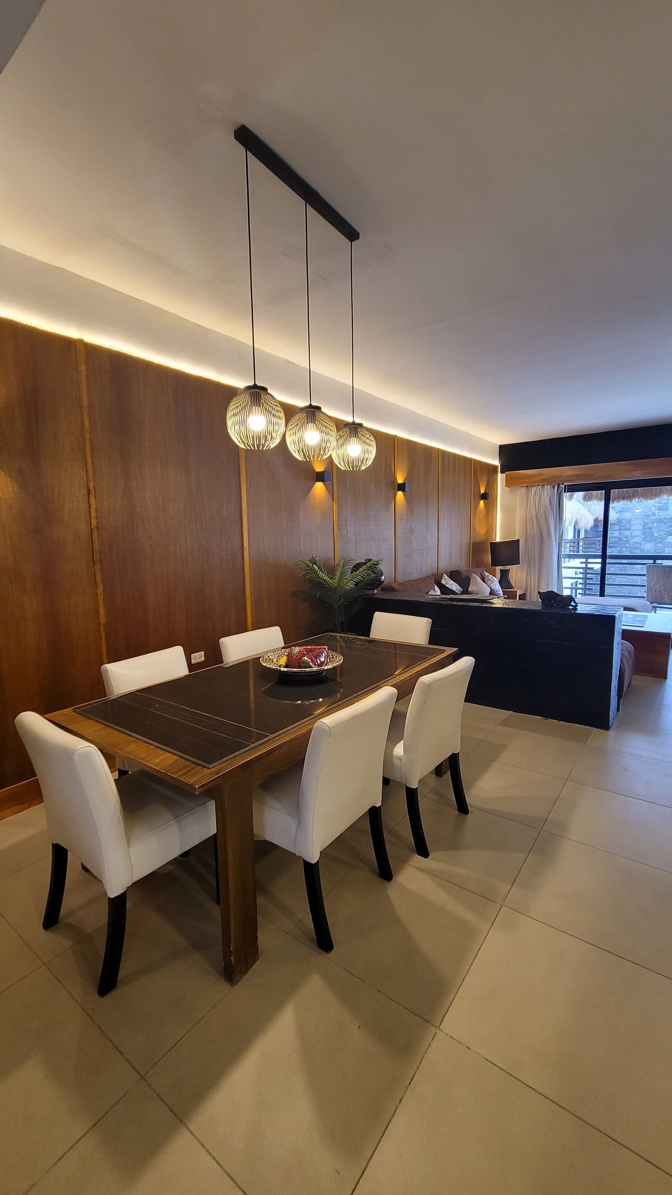 A modern dining area with a wooden table, white chairs, pendant lighting, and a view of the living area in the background.